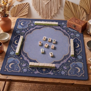 Può includere: Un tappetino da mahjong quadrato con un bordo blu scuro con motivi floreali e a mezzaluna, che circonda un'area centrale blu più chiara. Il tappetino è disposto con quattro file di tessere bianche e verdi e alcune tessere sparse al centro.