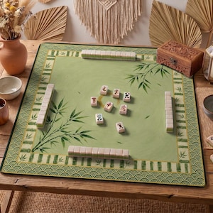 Könnte beinhalten: Eine grüne Mahjong-Spielmatte mit Bambusblatt-Design und dekorativem Rand. Die Matte liegt auf einem Holztisch mit Spielsteinen und einer Holzkiste. Das Spiel ist spielbereit.