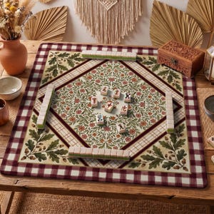 May include: A square mahjong mat featuring a central diamond pattern with floral motifs, surrounded by oak leaves and acorns. The border has a dark red and white checkered design. Several mahjong tiles are arranged on the mat surface.