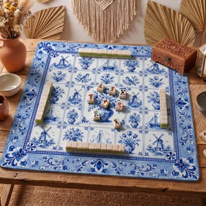 May include: A square game mat featuring a blue and white tile pattern with illustrations of windmills, sailboats, birds, and floral arrangements. The mat includes several mahjong tiles arranged in rows and scattered across the center surface.