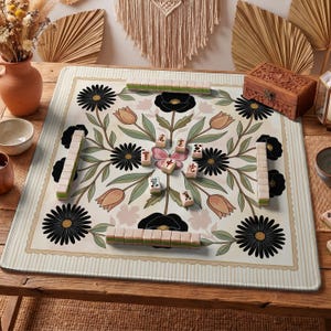 Puede incluir: Un juego de Mahjong sobre una alfombra crema con un diseño floral con margaritas negras, tulipanes rosas y hojas verdes. El juego incluye fichas y está dispuesto sobre una mesa de madera, con una caja de madera decorativa y otra decoración.