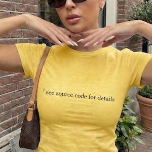 May include: A yellow short-sleeve t-shirt with the text "I see source code for details" in black. The person is wearing dark oval sunglasses and a small brown purse.