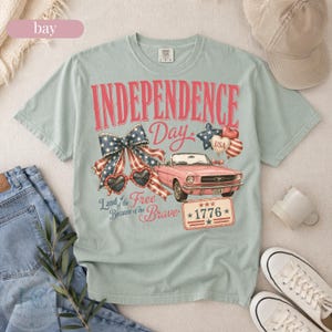 K&ouml;nnte beinhalten: Hellblaues T-Shirt mit einem Grafikdesign zum Unabh&auml;ngigkeitstag. Das Design enth&auml;lt die roten Worte "INDEPENDENCE DAY", ein rosa Cabrio und patriotische Elemente wie eine Schleife, Sonnenbrille und Luftballons.