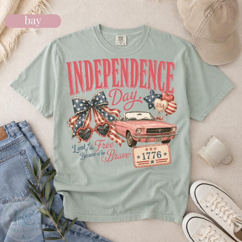 K&ouml;nnte beinhalten: Hellblaues T-Shirt mit einem Grafikdesign zum Unabh&auml;ngigkeitstag. Das Design enth&auml;lt die roten Worte "INDEPENDENCE DAY", ein rosa Cabrio und patriotische Elemente wie eine Schleife, Sonnenbrille und Luftballons.