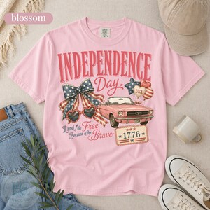 K&ouml;nnte beinhalten: Hellrosa Independence Day T-Shirt mit einem Aufdruck eines Oldtimer-Cabrios in Rosa, herzf&ouml;rmigen Sonnenbrillen und einer patriotischen Schleife. Das Shirt tr&auml;gt den Text "Independence Day" und "Land of the Free Because of the Brave."
