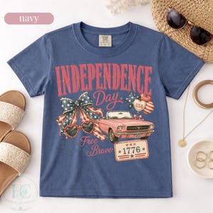 K&ouml;nnte beinhalten: Marineblaues T-Shirt mit einem Grafikdesign zum Unabh&auml;ngigkeitstag. Das Design zeigt ein rosa Cabrio, herzf&ouml;rmige Sonnenbrillen, eine Schleife und den Text "Independence Day", "Free Brave" und "1776".