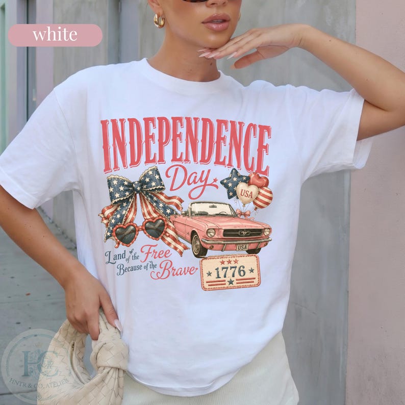 K&ouml;nnte beinhalten: Wei&szlig;es T-Shirt mit einem patriotischen Independence Day-Motiv. Das Design zeigt ein rosa Cabrio, herzf&ouml;rmige Sonnenbrillen, eine Schleife und den Text "INDEPENDENCE Day" und "Land of the Free Because of the Brave".