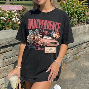 K&ouml;nnte beinhalten: Schwarzes T-Shirt mit einem patriotischen Independence Day-Motiv. Das Design zeigt ein rosa Auto, eine Schleife, herzf&ouml;rmige Sonnenbrillen und den Text "Independence Day" und "Land of the Free Because of the Brave" mit dem Jahr "1776".
