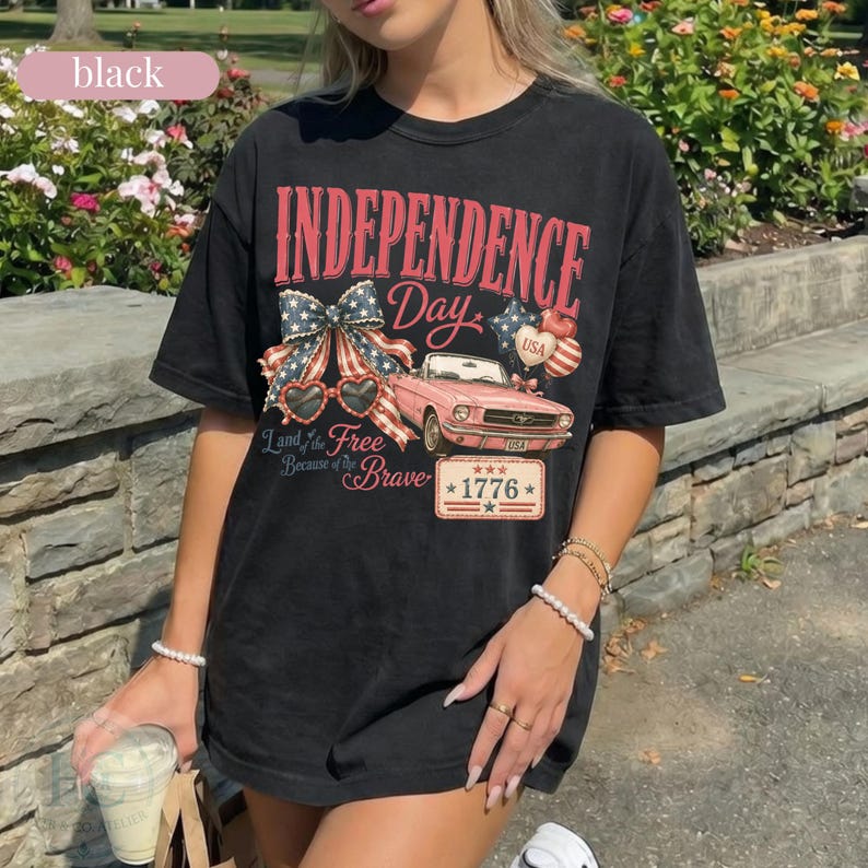 K&ouml;nnte beinhalten: Schwarzes T-Shirt mit einem patriotischen Independence Day-Motiv. Das Design zeigt ein rosa Auto, eine Schleife, herzf&ouml;rmige Sonnenbrillen und den Text "Independence Day" und "Land of the Free Because of the Brave" mit dem Jahr "1776".