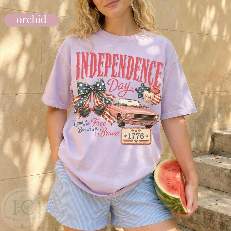 K&ouml;nnte beinhalten: Ein helllila Independence Day T-Shirt mit einer Grafik eines rosa Cabriolets, einer patriotischen Schleife, herzf&ouml;rmigen Sonnenbrillen und dem Text "INDEPENDENCE DAY 1776". Das Shirt hat auch den Text "Land of the Free Because of the Brave".