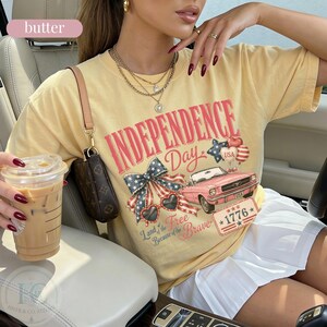 K&ouml;nnte beinhalten: Buttergelbes Independence Day T-Shirt mit einer Grafik eines rosa Autos, einer patriotischen Schleife und dem Text "Independence Day USA 1776". Das Shirt wird mit einem wei&szlig;en Rock getragen.