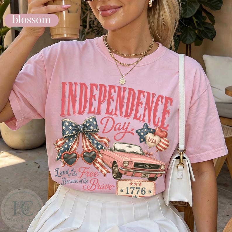 K&ouml;nnte beinhalten: Ein hellrosa T-Shirt mit den Worten "INDEPENDENCE Day" in Rot. Das Shirt zeigt ein patriotisches Design mit einer Schleife, herzf&ouml;rmigen Sonnenbrillen, einem rosa Auto und dem Text "Land of the Free Because of the Brave" und "1776".