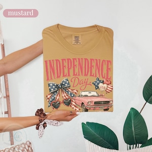 K&ouml;nnte beinhalten: Senffarbenes T-Shirt mit einem Grafikdesign zum Unabh&auml;ngigkeitstag. Das Design enth&auml;lt den Text "INDEPENDENCE Day", eine Schleife, herzf&ouml;rmige Sonnenbrillen, ein rosa Cabrio und Luftballons.