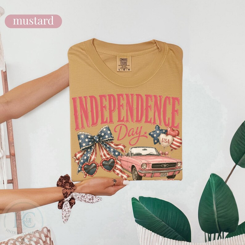 K&ouml;nnte beinhalten: Senffarbenes T-Shirt mit einem Grafikdesign zum Unabh&auml;ngigkeitstag. Das Design enth&auml;lt den Text "INDEPENDENCE Day", eine Schleife, herzf&ouml;rmige Sonnenbrillen, ein rosa Cabrio und Luftballons.