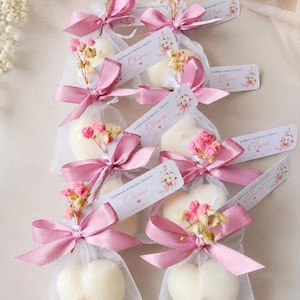 Op de afbeelding: Hartvormige, crèmekleurige zeep in transparante zakjes, vastgebonden met roze linten en versierd met gedroogde bloemen. Elk zakje heeft een label met bloemaccenten en de naam "Oliwia". Een delicaat cadeau.