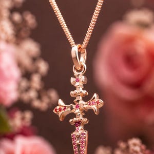 May include: A gold-colored pendant necklace featuring a cross-shaped design with embedded red gemstones. The pendant hangs from a delicate chain. The background is blurred, with pink and white flowers.