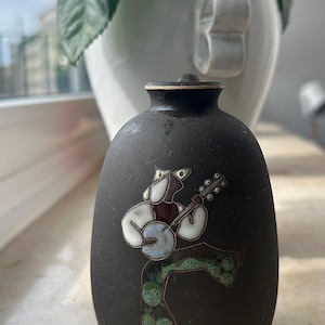 Könnte beinhalten: Kleine, schwarze Keramikvase mit rundem Körper und schmalem Hals. Sie zeigt eine farbenfrohe Illustration eines Banjospielers. Der Spieler trägt ein weißes Hemd, blaue Hosen und einen braunen Hut. Die Vase ist etwa 13 cm hoch.