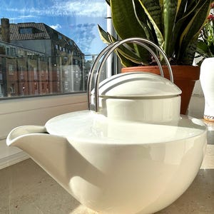 Op de afbeelding: Een witte keramische theepot met een glad, afgerond lichaam en een brede, geïntegreerde tuit. De pot heeft een gebogen handvat van gepolijst zilverkleurig metaal dat vastzit aan het deksel, dat naadloos in de ronde opening van de basis past.