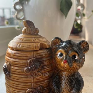 Könnte beinhalten: Ein Keramik-Honigtopf in Form eines Bienenstocks mit einer Bärenfigur. Der Honigtopf ist braun mit Bienen- und Blattdetails. Der Bär ist schwarz und braun mit einer roten Nase und sitzt neben dem Honigtopf.
