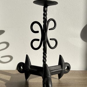 Op de afbeelding: Een zwarte metalen kandelaar met een driepotige basis met drie puntige voeten en een centrale verticale steel. De steel bevat een gedraaid metalen gedeelte en een decoratief krulmotief onder een ronde plaat met een centrale pin.