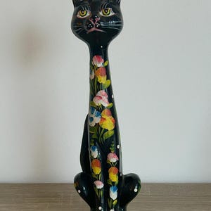 Könnte beinhalten: Eine hohe, schlanke schwarze Katzenfigur mit langem Hals und spitzen Ohren. Die Katze ist auf der Brust und den Vorderbeinen mit handgemalten Blumenmustern in Rosa, Gelb und Blau sowie kleinen weißen Punkten verziert.