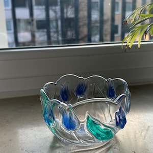 Könnte beinhalten: Eine klare Glasschale mit gewelltem Rand und farbigen Akzenten. Die Schale hat blaue und grüne blattartige Designs an den Rändern. Die Schale steht auf einer Steinoberfläche.