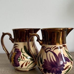 Op de afbeelding: Twee keramische kannen met een crèmekleurige basis, voorzien van handgeschilderde paarse bloemen en groene bladeren. De bovenranden en handvatten zijn afgewerkt met een metallic bronskleurige glazuurlaag die een reflecterend oppervlak biedt.