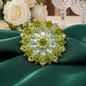 May include: A gold-toned, floral-shaped brooch with a central cluster of clear, faceted crystals surrounded by layers of green and chartreuse crystals. The brooch is set against a dark green satin fabric.