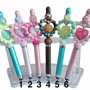 May include: A selection of six decorative stylus pens displayed in a clear stand. Each pen has a unique design, with heart-shaped frames, beaded details, and colourful tops. The pens come in various colours, including pink, teal, and brown.