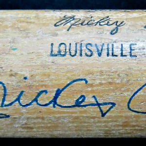 Pode incluir: Um bastão de beisebol de madeira com o texto "Mickey Mantle" e sua assinatura em tinta azul. O bastão também apresenta uma imagem pintada de Mickey Mantle em uniforme de beisebol e as palavras "Louisville Slugger".