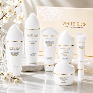 May include: A collection of white skincare products with gold accents, including toner, lotion, cleanser, serum, and cream. The packaging features the text "WHITE RICE ADD 15% NIACINAMIDE" and the brand name "SADOER". The products are arranged on a reflective surface.