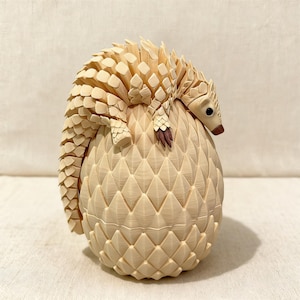 May include: A detailed, cream-colored pangolin sculpture. The pangolin is curled around a textured, egg-shaped form. The sculpture features intricate scales and a detailed head with a brown snout and small eyes. The overall design is artistic and decorative.
