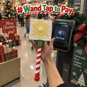 May include: A handheld wand featuring a white square top with a yellow snowflake design and a red and white candy cane striped handle. The wand is positioned in front of a black payment terminal with a screen displaying the text Tap to pay.