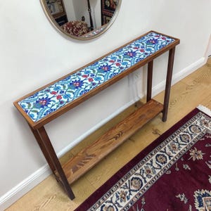 May include: A narrow, wooden console table with a tiled top featuring a floral pattern in blue, red, and white. The table has a lower shelf and is supported by angled legs. The table is approximately 48 inches long.