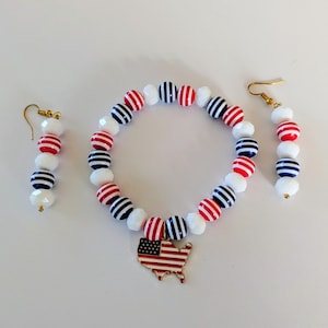May include: A jewelry set consisting of a beaded bracelet and two matching drop earrings. The beads are white, red-striped, and navy-striped. The bracelet features a gold-toned metal charm shaped like the United States map with flag colors.