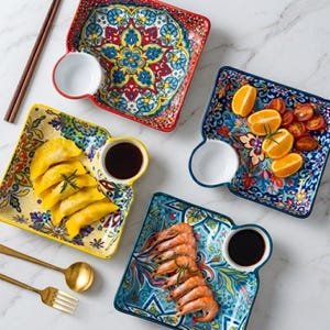 May include: A selection of square ceramic serving dishes with integrated sauce bowls. The plates, adorned with floral patterns in red, blue, yellow, and green, hold dumplings, shrimp, oranges, and tomatoes. Chopsticks and gold utensils are also visible. The dishes are ideal for serving appetizers.