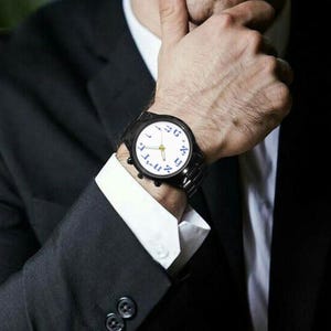 May include: A black wristwatch with a white face and blue numerals is displayed. The watch has a black band and is worn on a wrist. The person is wearing a black suit with a white collared shirt.