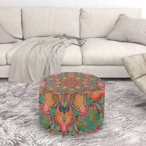 May include: A round ottoman with a colorful, intricate mandala design in shades of orange, teal, and pink. The ottoman is placed on a fluffy, white rug in front of a white sofa.