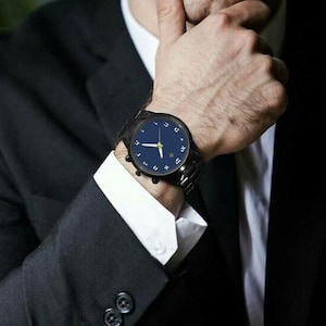 May include: A black wristwatch with a dark blue face and Arabic numerals. The watch has a black metal band and is being worn on a wrist. The watch hands are gold. The person is wearing a black suit with a white dress shirt.
