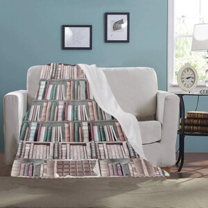 Pode incluir: Uma manta de l&atilde; branca e macia com um design estampado de uma estante cheia de livros coloridos. A estante &eacute; de madeira e tem uma barra de chocolate na prateleira inferior.