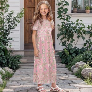 Può includere: Una giovane persona indossa un vestito rosa con stampa floreale e sandali bianchi. L'abito presenta un motivo ripetuto di fiori e foglie. La persona è in piedi su un sentiero di pietra di fronte a una casa.