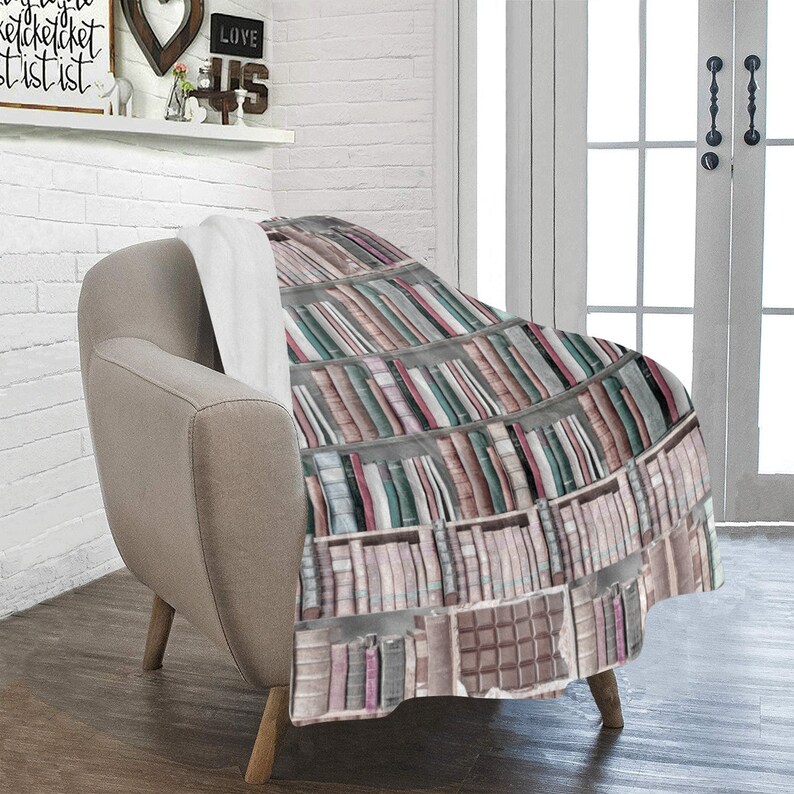 Pode incluir: Uma manta macia e felpuda com um design de estante de livros que apresenta fileiras de livros coloridos. A manta est&aacute; drapeada sobre uma poltrona marrom clara.