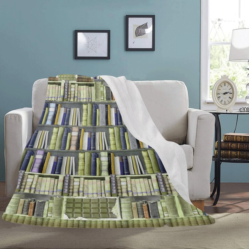 Pode incluir: Uma manta branca com um padr&atilde;o de estante de livros impresso. A estante est&aacute; cheia de livros coloridos. A manta est&aacute; colocada sobre um sof&aacute; branco.