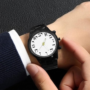 May include: Black wristwatch with a white face featuring Hebrew numerals. The watch has a black metal band and a black case. The hands are gold-colored. The watch is being worn on a wrist.