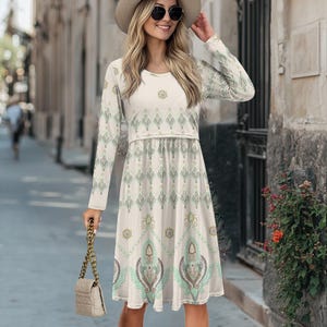 Könnte beinhalten: Ein weißes, langärmeliges Kleid mit einem geometrischen Muster in Grün und Gold. Das Kleid hat einen runden Ausschnitt und einen fließenden Rock.