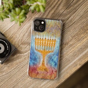 May include: Clear phone case featuring a colorful design of a golden menorah. The background has a textured, abstract appearance with shades of blue, gold, and red. The phone case is on a wooden surface.