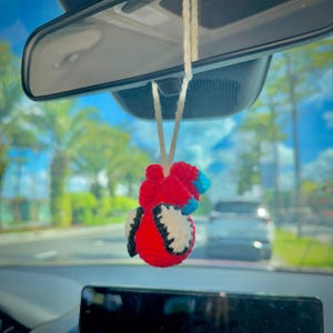 May include: A crocheted hanging ornament featuring a red heart-shaped design with blue accents and two white wing-like appendages with black outlines, suspended from a white string attached to a vehicle rearview mirror.