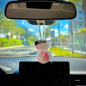 May include: A small, handmade crochet ornament hanging from a rearview mirror. The figure is primarily white with black accents on the top and pink details on the bottom, featuring a rounded shape suspended by a white yarn loop for display.