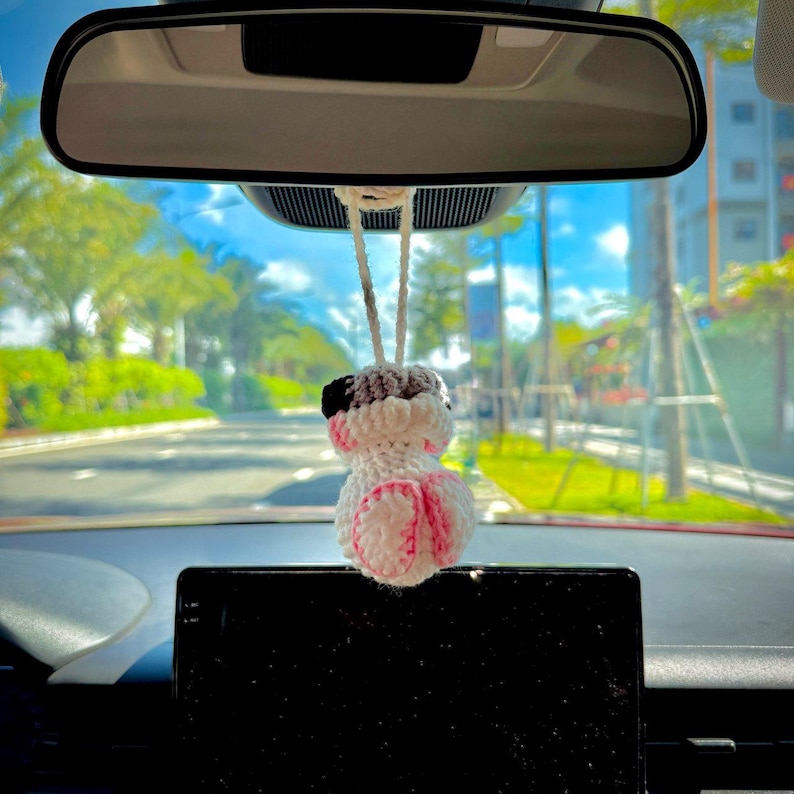 May include: A small, handmade crochet ornament hanging from a rearview mirror. The figure is primarily white with black accents on the top and pink details on the bottom, featuring a rounded shape suspended by a white yarn loop for display.