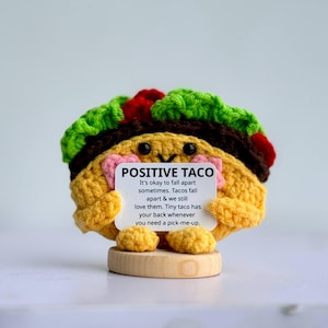May include: A crocheted taco figurine with a smiling face, holding a sign that reads "POSITIVE TACO." The taco has green, red, and brown toppings. It sits on a small wooden base. The sign offers words of encouragement.